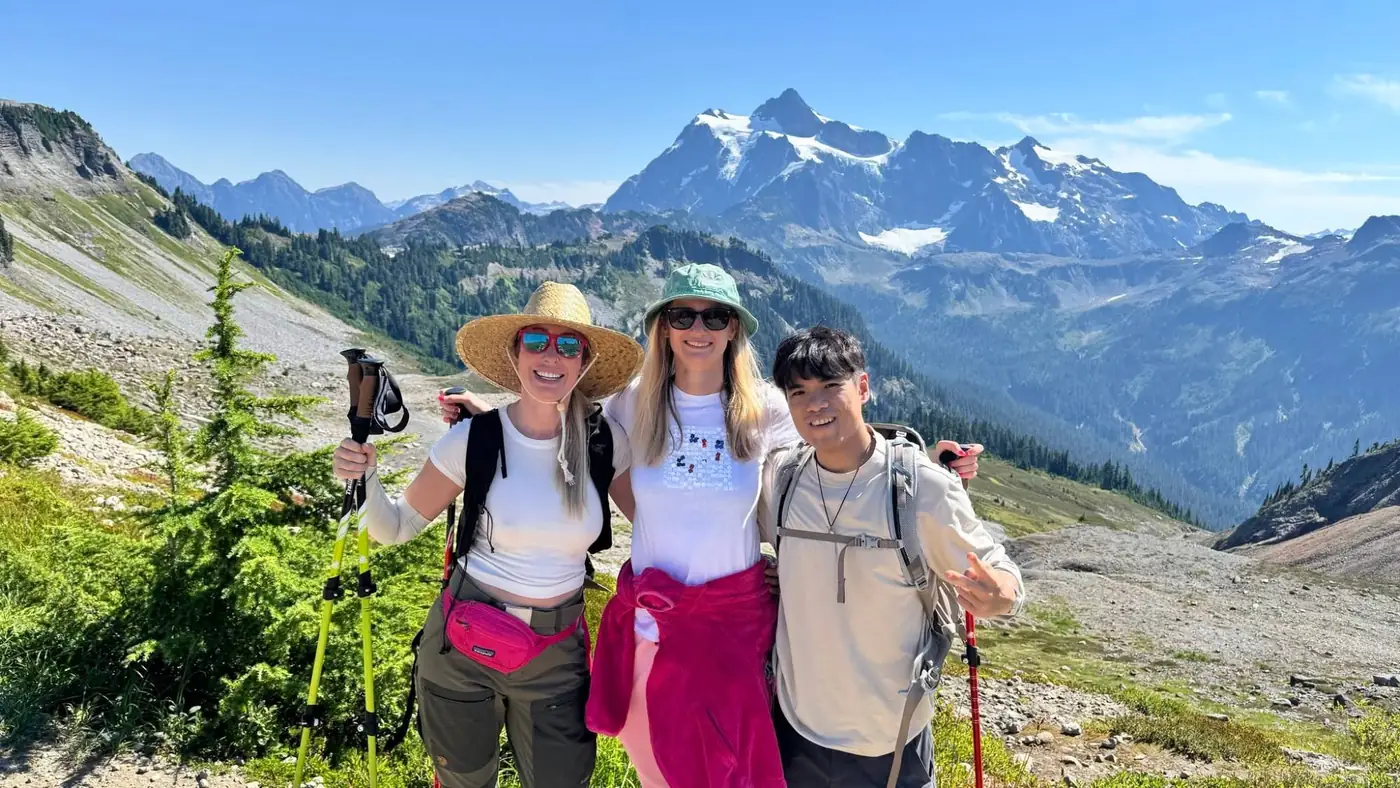 Three hikers smiling on an alpine trail with a snow-capped mountain behind them